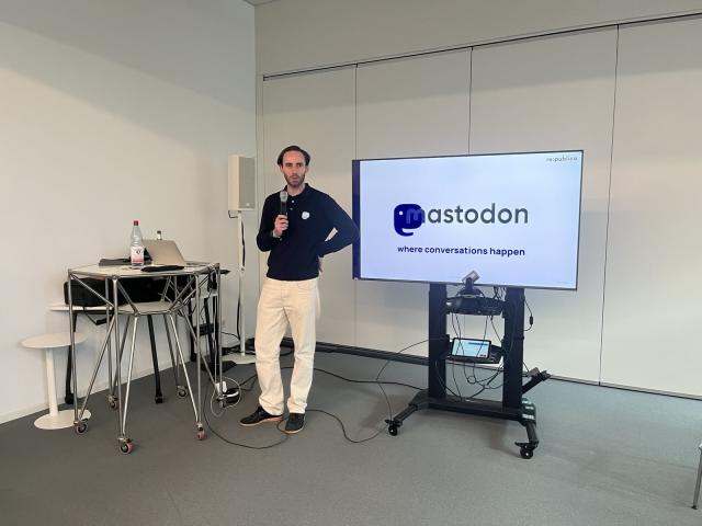 EIne Person steht mit einem Mikro neben einem Bildschirm. Auf dem Bildschirm ist das Logo Mastodon von Mastodon zu sehen. Links neben dem Mann steht ein Tisch. 
