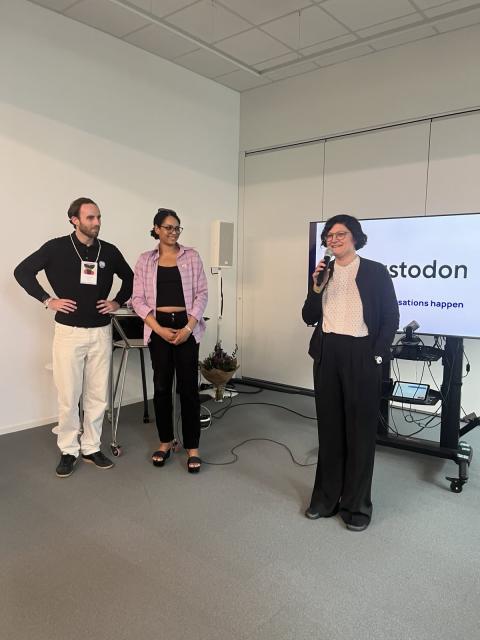 Franziska Heine, Geschäftsführende Vorständin von Wikimedia Deutschland e. V. steht mit einem Mikrofon vor dem Bildschirm ist das Logo Mastodo. Links daneben stehen zwei Personen von Mastodon, ein Mann und eine Frau. 