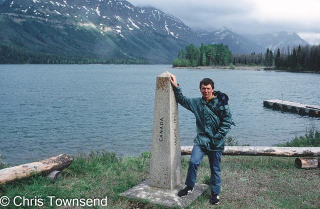 The poster in a rain jacket standing by an obelisk marking the US/Canada border beside a lake with snow-spattered mountains rising into clouds in the distance.