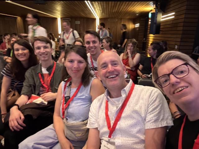 5 humans sitting together and smiling, all wearing red lanyards