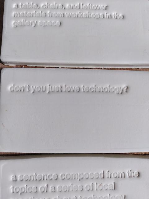 three porcelain subway tiles, with the texts "a table, chairs and leftover materials from workshops in the gallery space", "don't you just love technology?", and "a sentence composed from the topics of a series of local meetups about technology"