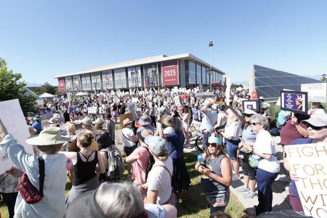 A wide shot at the Marriott Library plaza on the University of Utah campus. Hundreds of people can be seen in the shot, many holding up political signs protesting Donald Trump.
