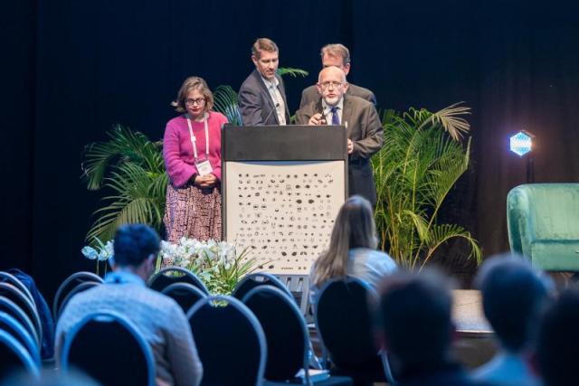 Four people stand around a podium at a conference; the podium has an artwork on it, showing hundreds of sets of eyes.