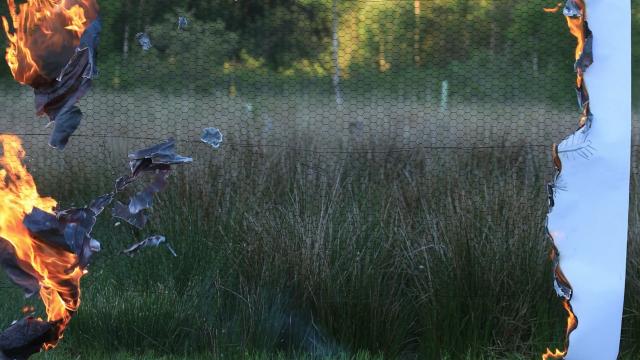 Paper burning against a fence in a field, with a large gap in the middle where you can see a meadow in the background