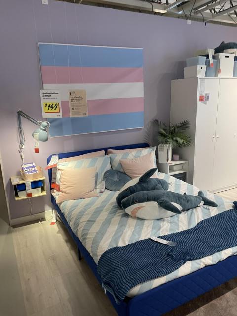 A photograph of a display bed in IKEA with two blahajs on it. Behind it on the wall is a trans flag