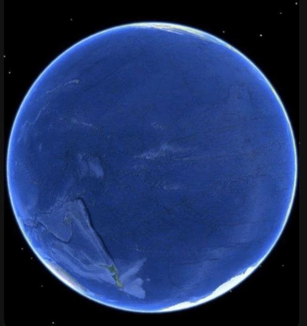 Satellite view of Earth showing primarily the Pacific Ocean side, with minimal visible landmasses. The image shows Earth as a blue sphere against the black of space, with a bright rim of atmospheric glow along the edge. This perspective showcases the vast expanse of the Pacific Ocean, which dominates most of the visible hemisphere, with only small portions of land visible near the edges, likely parts of Antarctica and South America. The ocean appears as various shades of deep blue, with some cloud patterns visible across its surface.