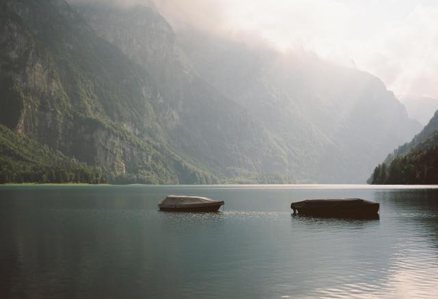 The picture shows a mountain lake surrounded by tree-covered rocks. Two boats are floating near the shore. The sun is shining onto the lake and the trees through a layer of clouds giving the scene a warm and calm feeling.