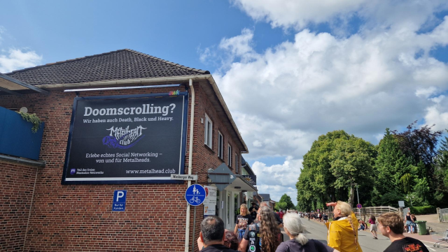 Our "Doomscrolling? We also have Death, Black and Heavy" poster on a brick wall in Wacken. People standing in front of it are looking at it.