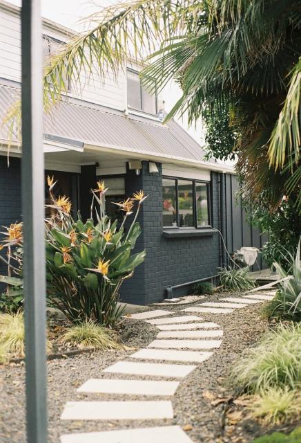 A footpath made from pavers and stones weaving through a garden toward a denim-blue two-storey house with palm trees.