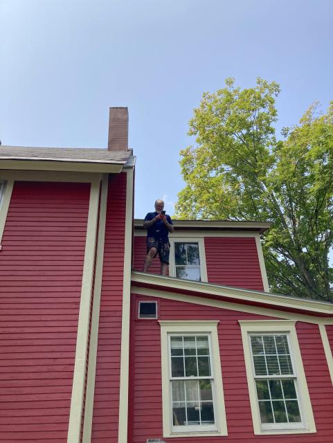 A man wearing reading glasses looks at his phone from the sloping porch roof of a red house with pale yellow trim