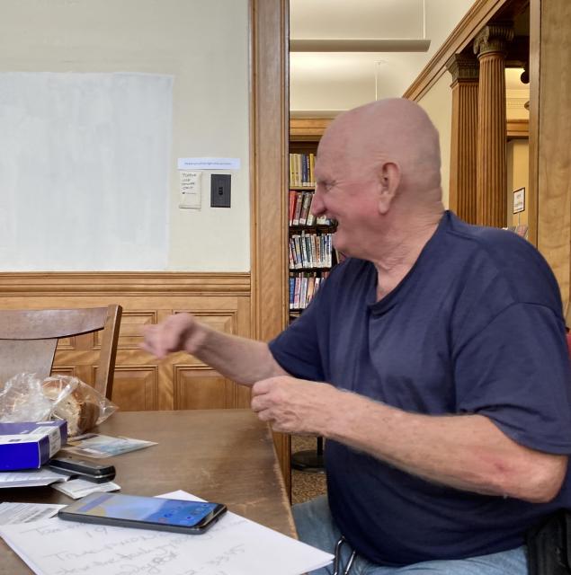 An older heavyset bald white man in a blue t-shirt with a pile pf papers and an android phone in front of him, waves his hands