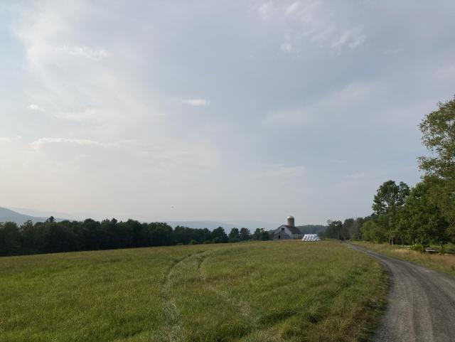 A long view down a dirt road showing a white barn with some hay bales next to it, a field in the foreground, trees in the middle distance and some hills in the back. The sky is a hazy blue and there are no other people around