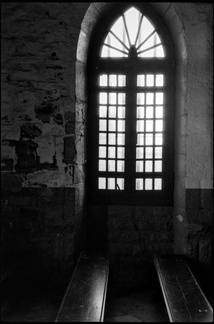 Black and white film photograph from inside a stone chapel, focused on a tall gothic window, with two pews below extending towards the camera, and the interior walls horizontally bisected by light and dark paint colors.