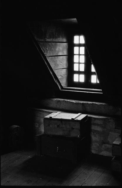 Black and white film photograph of an interior room, looking towards a gabled window, with wooded floors, and wood and stone walls, and a few wooden crates at the base of the window cove.