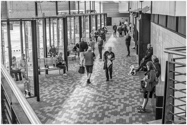A black-and-white image of a busy bus station with people sitting and standing in a waiting area. Several individuals are seated on benches while others are walking or waiting around. The floor features a patterned tile design, and glass panels create a modern atmosphere.