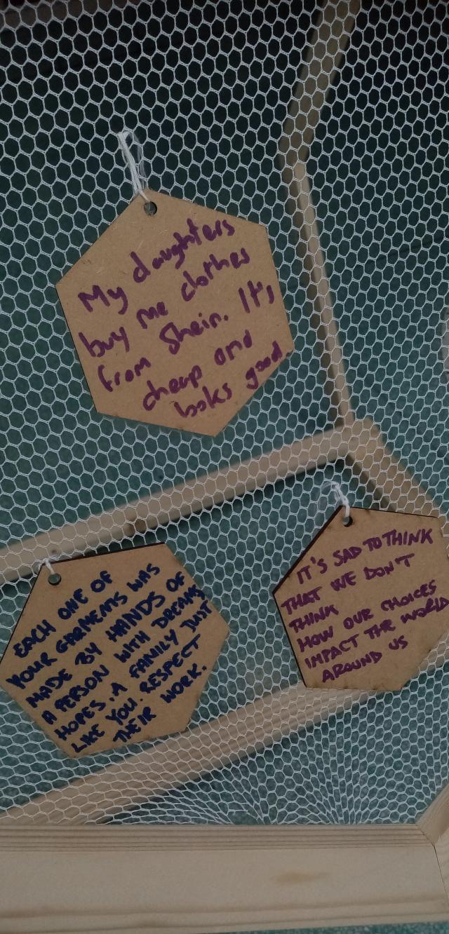 Three wooden hexagons hanging from a fabric tulle, each with a comment about clothing written in marker. One compliments the online retail space shein while the other teo ask for respect of those who make our clothes