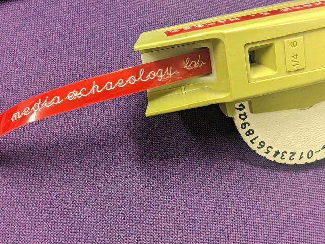 pale green Dymo embossing labeler, with a cursive embossing wheel. red tape reads "media archaeology lab"