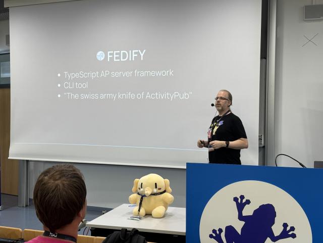 David Roetzel, a white man wearing a black t-shirt in front of a slide describing Fedify (a TypeScript ActivityPub server framework and CLI tool). A Mastodon plushie sits on the table in front.