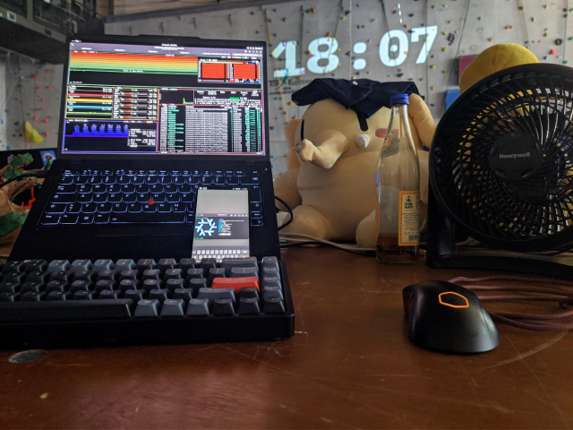 an image of the abenteuerhalle kalk, featuring the climbing wall with the evoke clock, a laptop, a phone running nixos, a laptop compiling at 100% cpu and most importantly the mastodon plush. also a fan which is very nice otherwise it'd just fucking melt