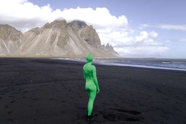 A person in a green morphsuit is standing with their back to the photographer, looking at the sea, a black sand beach, and a mountain in front of them. The mountain is called Vestrahorn, and it’s located in southeast Iceland.