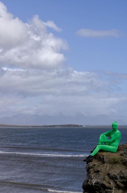 The same person in a morphsuit is sitting on a large rock, facing the camera. Behind them is a relatively calm sea.