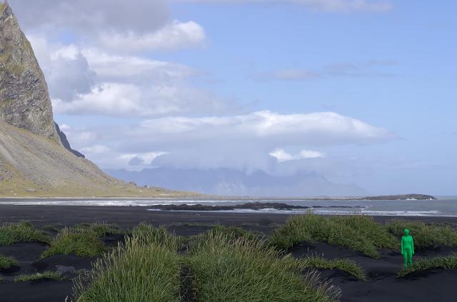 In the bottom right corner of the photo, the same person is standing on a black sand beach, quite far from the sea. Mountains are in the background, and large patches of marram grass (or some other tall, wetland-like grass) grow in the sand.