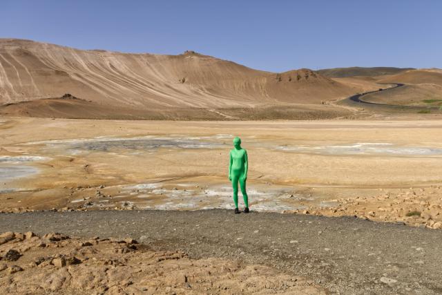 A geothermal area near Mývatn in Iceland (Hverir). The photo is almost entirely filled with orange and brown hues because of the mountains, rocks, and mud, with blue sky and gray sulphurous mud springs. The same green person is standing in the middle of the image, looking slightly away from the photographer.