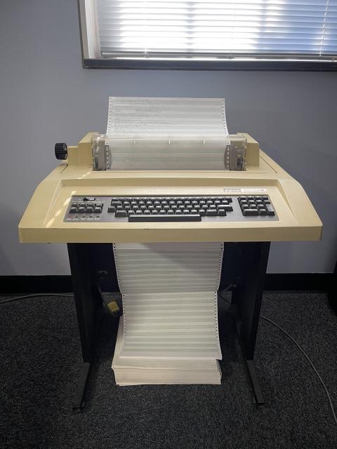 A Decwriter ii hardcopy terminal next to a wall and below an office window. Wide format greenbar paper is being fed through the machine. 