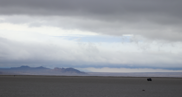 a flat expanse of grey is seen under a cloudy sky with different shades of grey clouds. a line of dark blue mountains rises to the left on the horizon in the far off distance