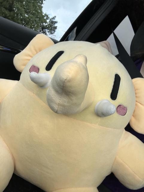 A large, plush yellow mastodon toy with black eyes and a small trunk is situated inside a vehicle. The scene features the plush toy in focus, while parts of the car interior and a blurred outdoor background are visible.