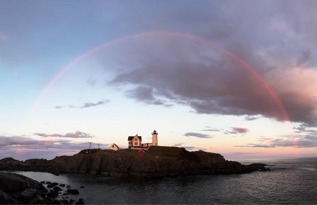 Lighthouse on a rocky peninsula at dusk surrounded by a 180 degree arc of a rainbow. (Taken with a phone in panorama mode; tone curve, perspective correction, and selective sharpening in Affinity Photo. Not generated by AI, no sky replacement, etc.)