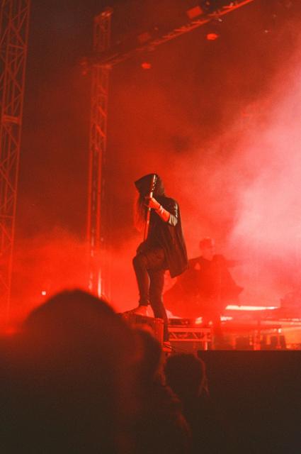 Amos Williams playing bass guitar, dressed in a hoodie, standing at the front of the stage with one leg on an elevation, illuminated by red light.