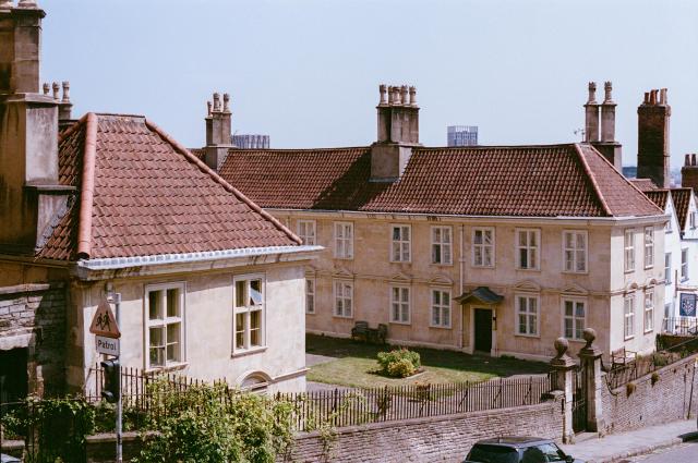An expansive beige two-story building with white window frames, a red tile roof and multiple chimneys, surrounded by a stone wall on the side of a steep road.