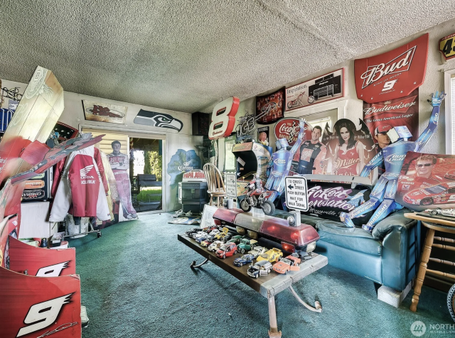 A very shabby living room absolutely covered by memorabilia for car racing and Budweiser beer. There is also a large set of police car lights taking up most of the coffee table. Every surface is just cluttered. 