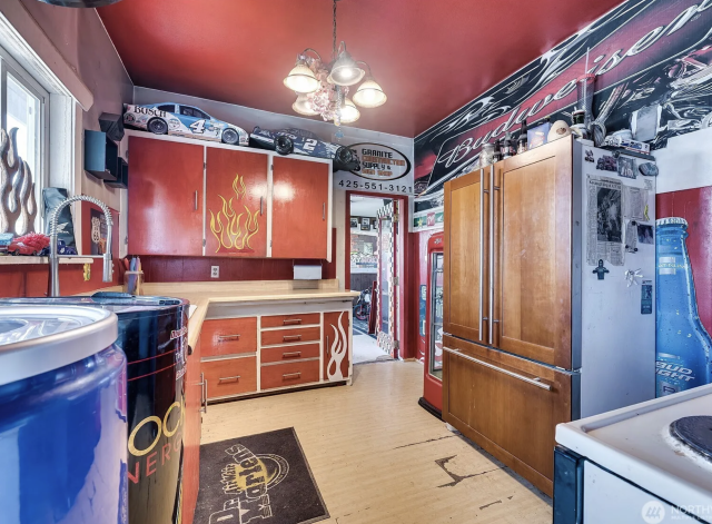 A kitchen with a red ceiling and red cabinets, with fake flames on the cabinet and a Budweiser logo across the wall, along with a blue beer bottle image on the wall, and 2 coolers for energy drinks. There are also model racing cars above the cabinets. 