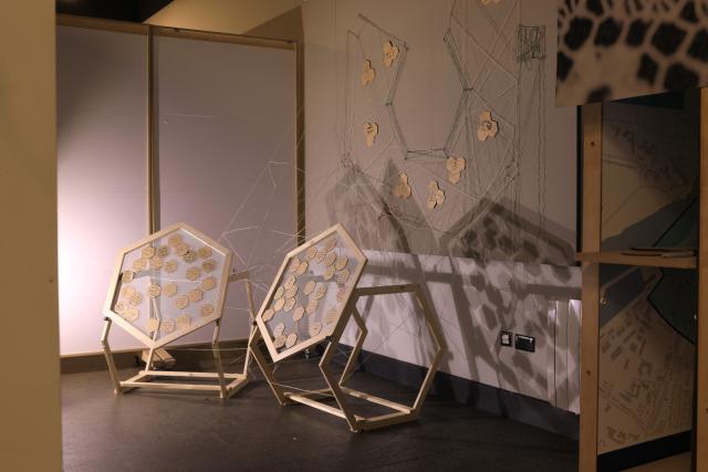 An art installation in a room lit from below, with two hexagonal timber sculptural pieces connected by string to a string drawing on the wall behind