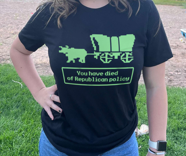 A t-shirt with an image ein the pixellated style of the old Oregon Trail game. A covered wagon pulled by a cow with the caption beneath it: 
You have died of Republican Policy