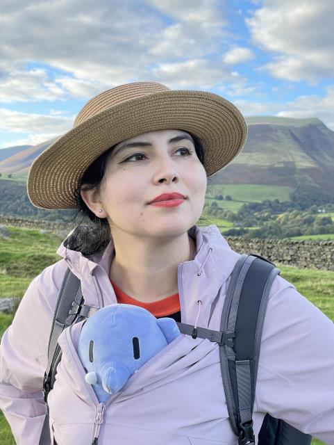 My wife dressed in outdoor clothing with a blue stuffed Mastodon toy peaking out from her jacket in front of a lusciously green landscape. 