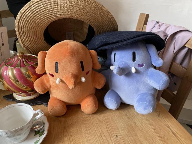 An apricot colored and a blue Mastodon stuffed toys sitting on a table next to each other. The blue one is wearing a human sized blue hat, and the apricot one a straw hat. 