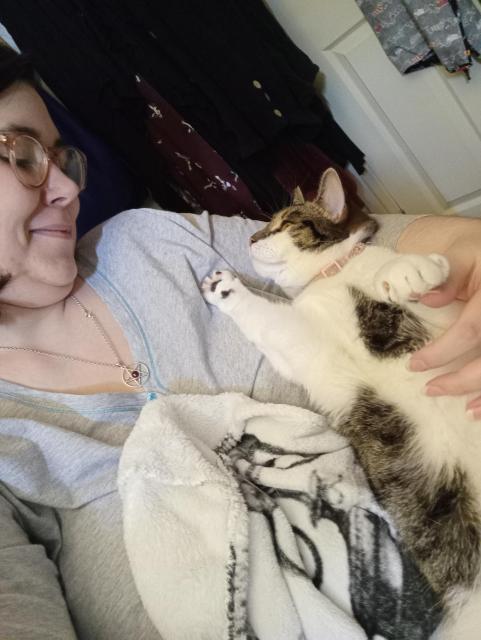 Me, a white enby with glasses, smiling as she peers up at me from the crook of my arm whilst making biscuits in the air.