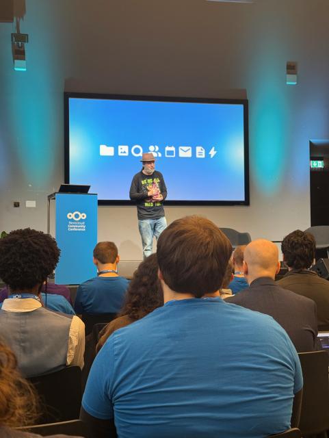 Jos Poortvliet, Director Communications & Co-Founder von Nextcloud, steht auf der Bühne der Nextcloud Community Conference. Er trägt einen Hut, ein schwarzes Shirt mit grün-gelber Aufschrift „We’re all mad here“ und eine bunte Stoffkatze in den Händen. Hinter ihm ist eine Leinwand mit Symbolen für Dateien, Suche, Kontakte, Kalender, Mail und weitere Funktionen zu sehen. Im Vordergrund sitzt das Publikum, viele in blauen Nextcloud-T-Shirts.