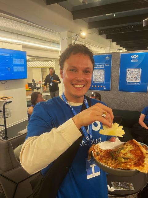 Someone from the Nextcloud team holding a plate of pizza in one hand and a slice of pineapple in the other hand, smiling at the camera.