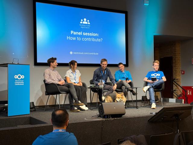 Fünf Personen sitzen auf einer Bühne bei der Nextcloud Community Conference während einer Paneldiskussion zum Thema ‚How to contribute?‘. Von links nach rechts: Barthélémy Briand, Dorra Jaouad, Simon Lindner, Anna Larch und Andrew Backhouse. Im Hintergrund ist eine große Leinwand mit der Aufschrift ‚Panel session: How to contribute?‘ und dem Nextcloud-Link zu sehen. Vor den Panelisten sitzt ein Hund am Bühnenrand.