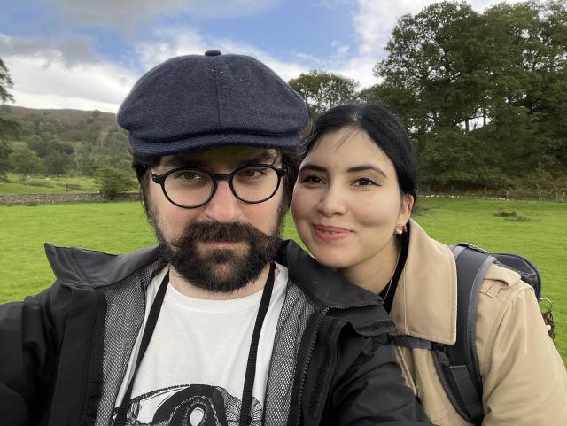 A man in a blue tweed cap with a somewhat curly thick mustache and black rimmed round glasses, wearing a rain jacket and a white Papangu band t-shirt, with a camera strap around his neck, next to a beautiful woman with dark hair, wearing a beige trench coat and a backpack. In the background, green grass and woods as well as a dry-stone wall.