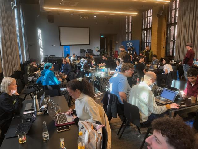 A picture of the main workspace at bUm. There are various tables with people working on laptops. Some people are standing and talking.