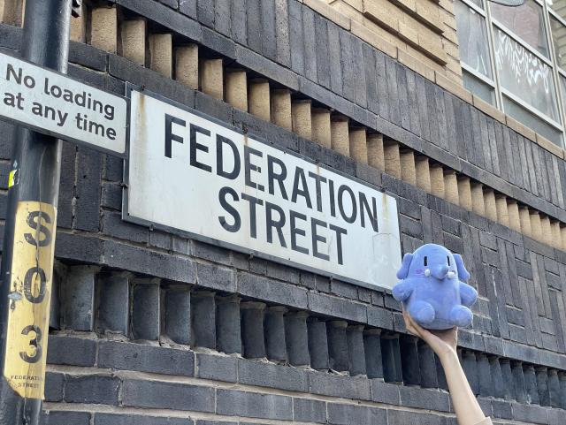 A little blue stuffed Mastodon toy being held up by one hand in front of the Federation Street sign. 