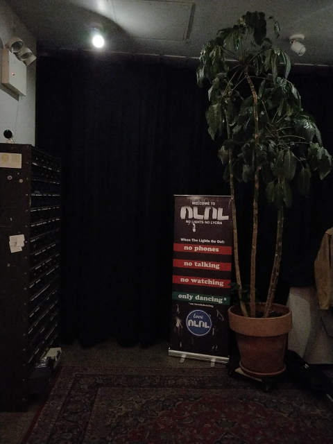 A black curtain flanked by a cabinet, plant, and a sign that says: Welcome to No Lights No Lycra. When the lights go out: no phones, no talking, no watching, only dancing* (*no break dancing)