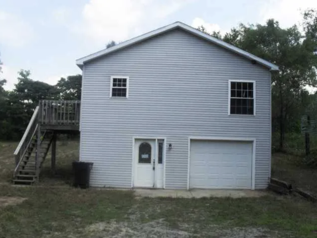 A grey house with a garage, a front door and only 2 windows. One window is small and one is big and they don't line up. It is a 2 story house and there is a lot of empty wall space where more windows should be. 