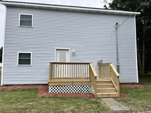 A grey 2 story house with timber steps up to a door, and only 2 small windows off to one side. There is also a lot of empty space where windows should go. 