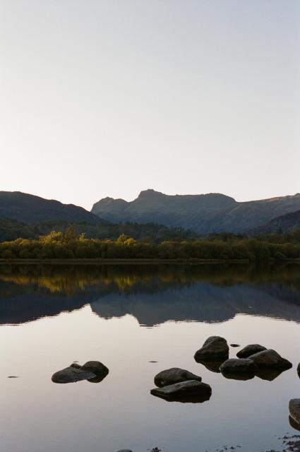 A calm lake, reflecting the auburn trees on the far shore and the Langdale pikes in the distance. Some rocks are in the water.